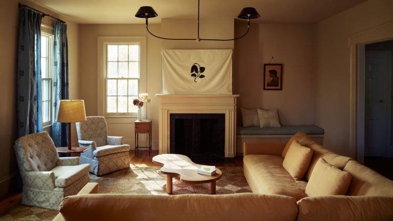 L’inspiration derrière cette maison de la vallée de l’Hudson est venue d’une source très inattendue : un naufrage tragique du XIXe siècle et une veuve en deuil Cozy living room with a cream fireplace, a unique organic-shaped wood coffee table, two patterned armchairs, and a large white banner with a black leaf design over the mantel