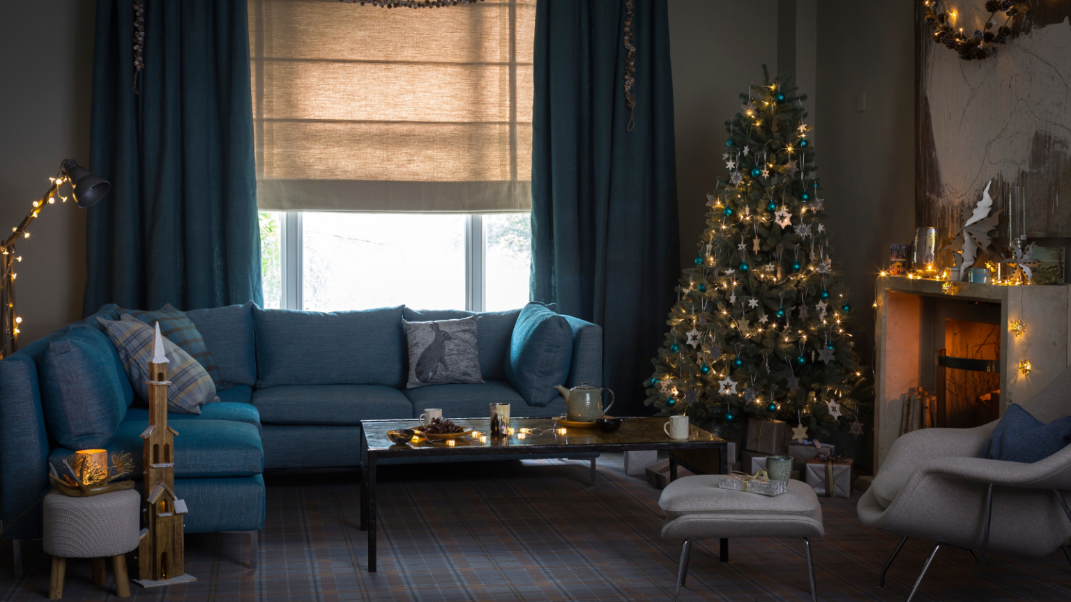 5 travaux essentiels d’entretien hivernal de la maison que les experts exhortent les propriétaires à faire avant le 30 novembre – Évitez les risques d’incendie, les robinets gelés et les réparations coûteuses A cozy, festive blue living room with a large blue sofa, decorated Christmas tree and white mantelpiece with a lit fire. In the background is a large window with neutral blinds and blue curtains, and in the foreground is a matching white chair with footrest.