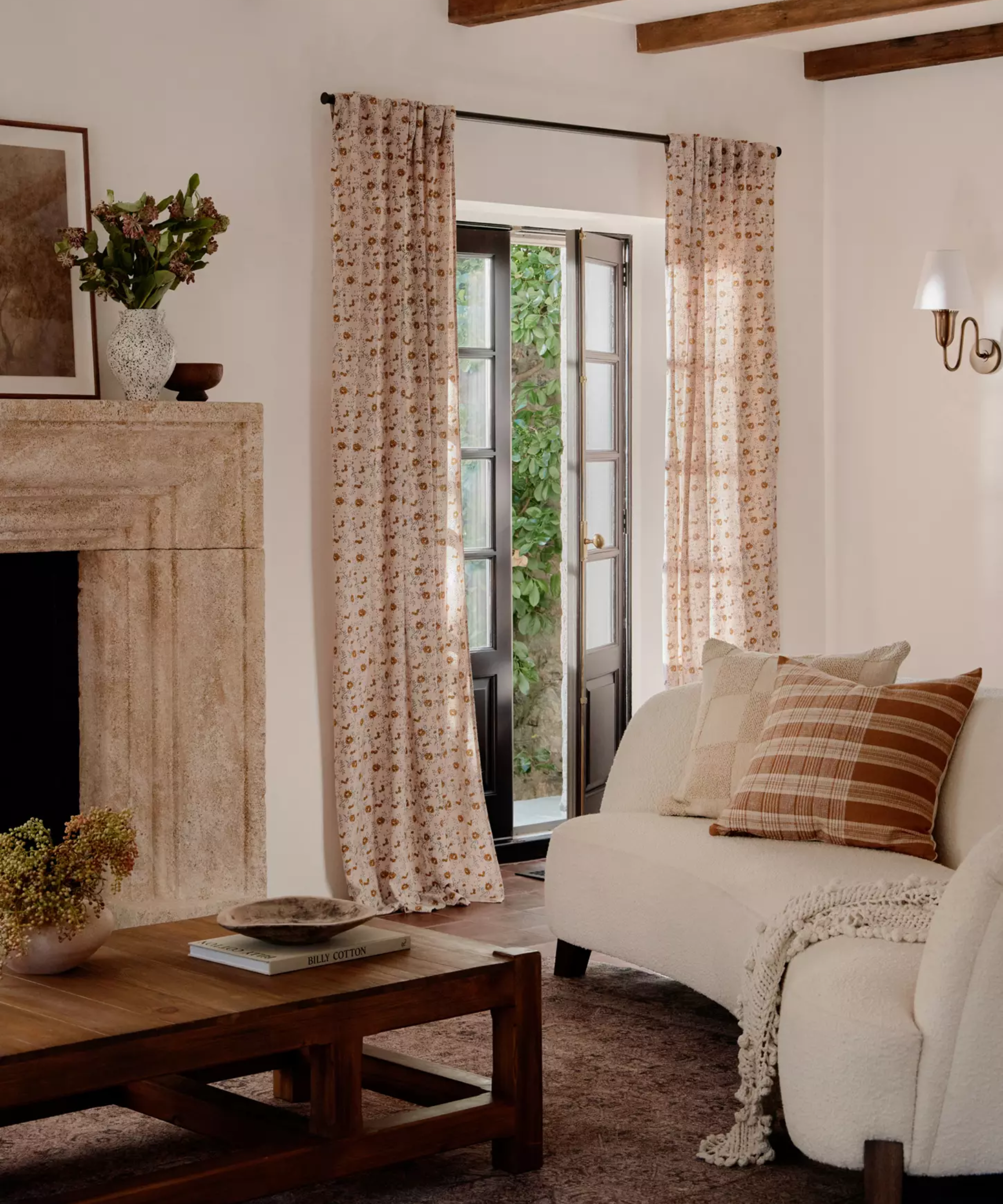 un salon avec de longs rideaux fleuris au-dessus des portes françaises, un canapé boucle, des oreillers, une table basse en bois et une cheminée