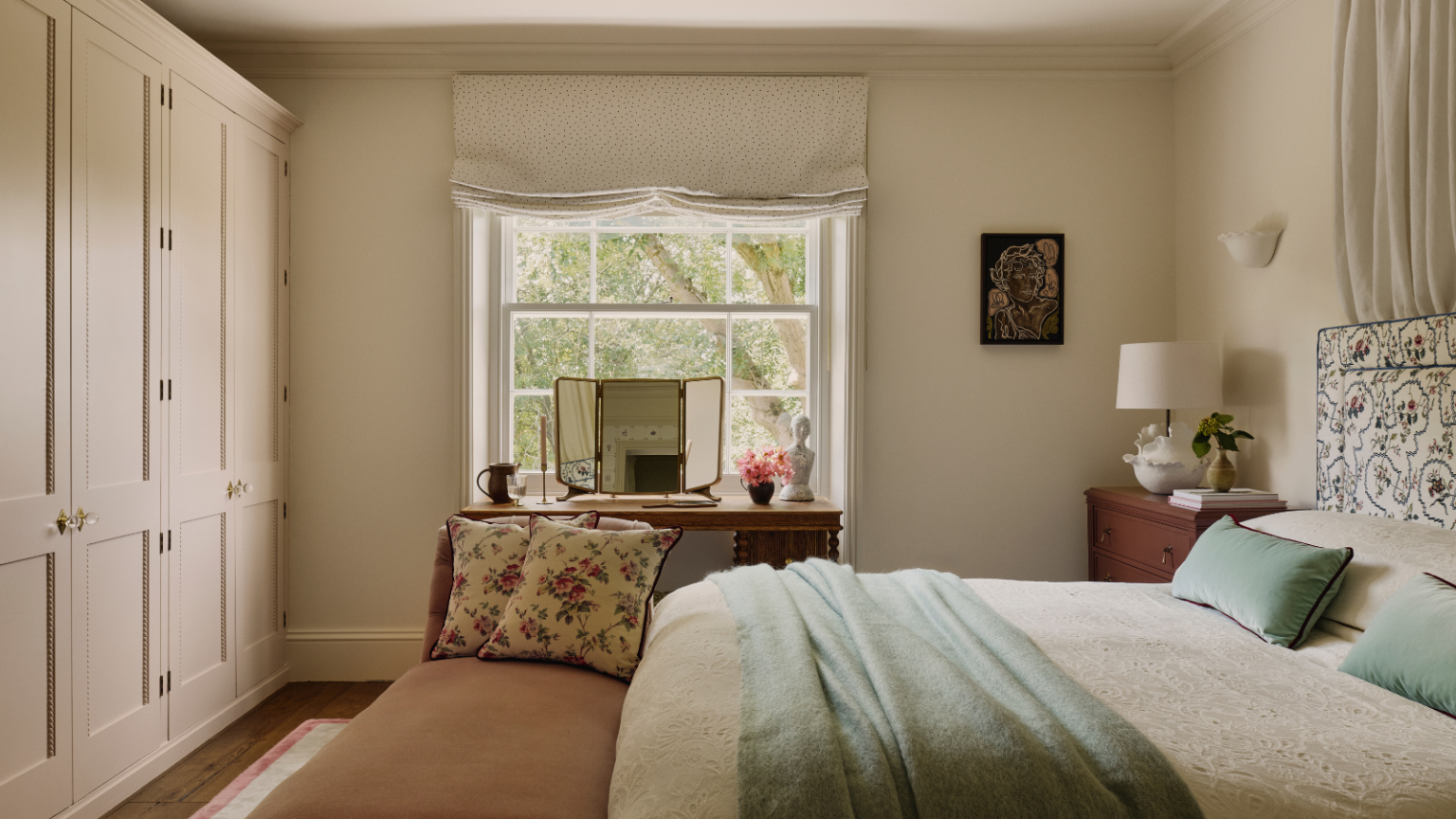Je transforme ma chambre d’amis en une suite d’invités 5 étoiles cette saison des fêtes – et ce porte-bagages Anthropologie est la clé pour y parvenir a bedroom with neutral colored walls, a wall of closets, a large window, a bed with cream colored sheets, a teal blanket, and teal pillows, and a bench at the foot of the bed with two floral pillows
