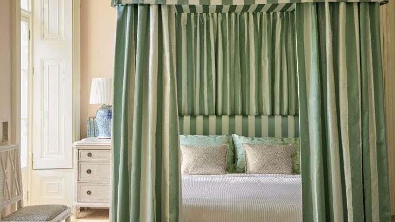 J’ai essayé la règle de désencombrement 90/90, et sa simplicité m’a fait aimer le minimalisme – cette méthode simple est idéale pour les invités. Cream bedroom with four-poster frame and striped green and cream curtains, and matching headboard