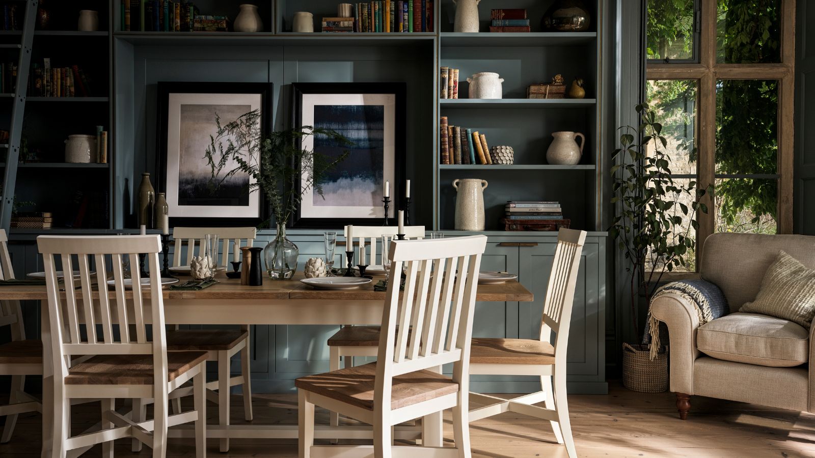 Une salle à manger avec de grandes étagères intégrées vert foncé sur le mur du fond, à côté d'une fenêtre. Un fauteuil rembourré à côté. Une table à manger rectangulaire blanche et en bois devant et des chaises assorties qui l'entourent. 