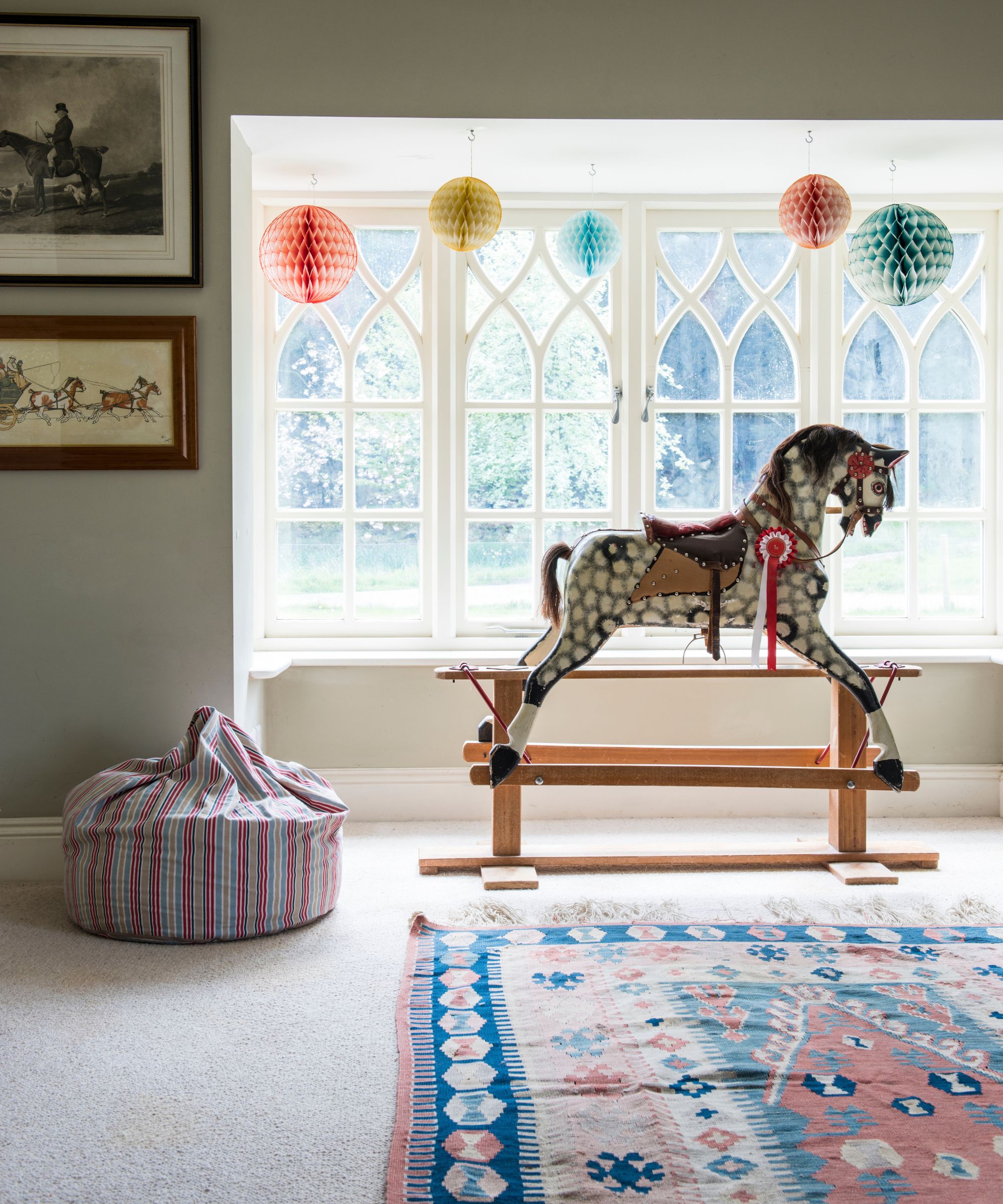 Salle de jeux avec tapis à motifs, cheval à bascule devant une grande fenêtre, pouf rayé et œuvres d'art représentant des chevaux.