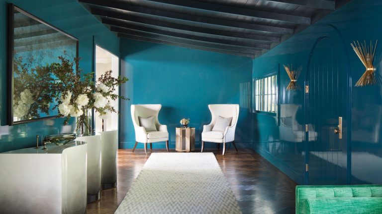 Room with sleek blue walls, two white armchairs in background, a white rug runner, marble-esque shelf with ornaments and flowers to the left.