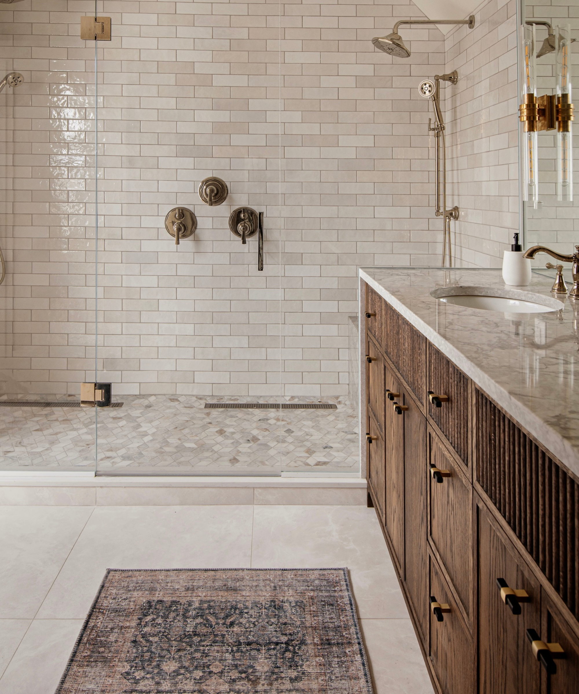 une salle de bain avec des murs carrelés blanc cassé dans la douche, un comptoir en marbre beige pour la vanité, des armoires en bois sombre en dessous et un tapis persan au sol