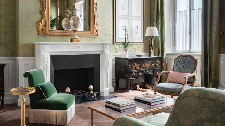 A traditional living room with green wallpaper, a large fireplace, a glass coffee table, and green armchairs.