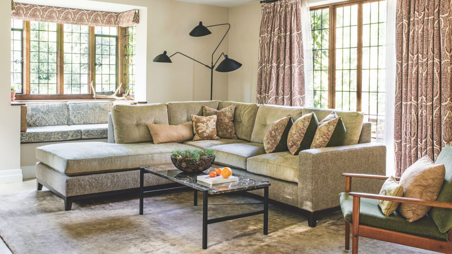 Des experts en fenêtres révèlent comment mettre la vôtre en « mode hiver » ne prend que 10 secondes et réduit la perte de chaleur jusqu’à un tiers A cozy living room with a large L-shaped velvet couch with cushions, a large velvet rug, marble coffee table, and large windows with patterned curtains. In the back corner is a large black lamp, and in the foreground is a green chair with a wooden frame and two cushions.