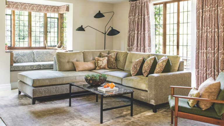 A cozy living room with a large L-shaped velvet couch with cushions, a large velvet rug, marble coffee table, and large windows with patterned curtains. In the back corner is a large black lamp, and in the foreground is a green chair with a wooden frame and two cushions.