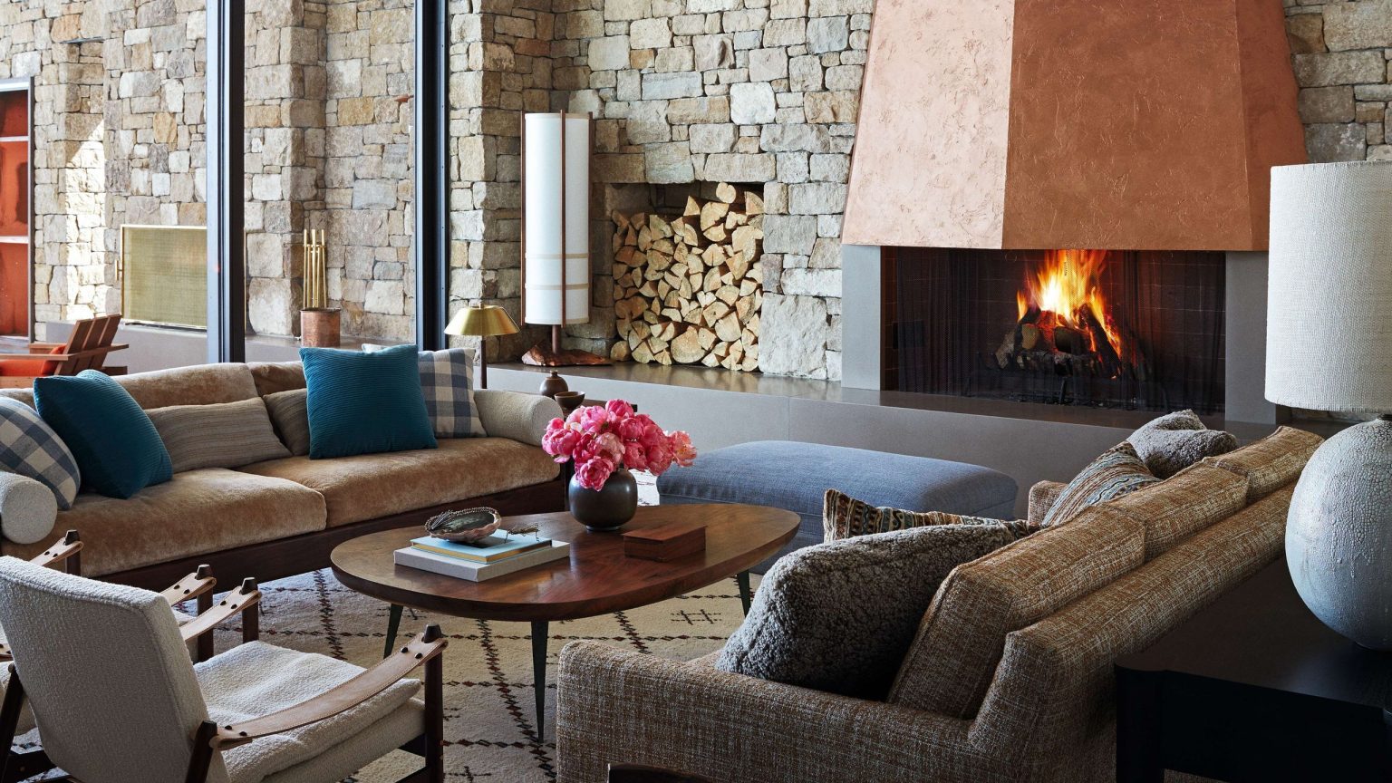 Il s’agit d’un chalet de ski, mais sans aucun décor de chalet de ski – Explorez une escapade à la montagne détendue, raffinée et prête à se divertir. A spacious, rustic living room featuring a towering copper fireplace, stone walls, large windows, and contemporary neutral-toned furniture.