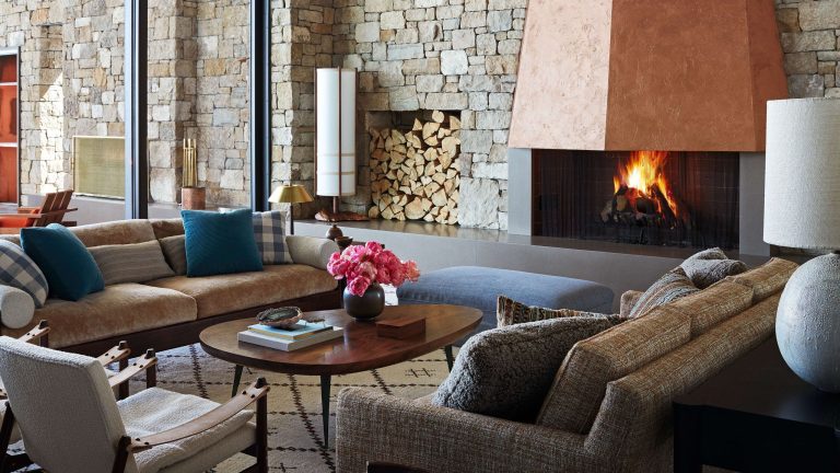 A spacious, rustic living room featuring a towering copper fireplace, stone walls, large windows, and contemporary neutral-toned furniture.