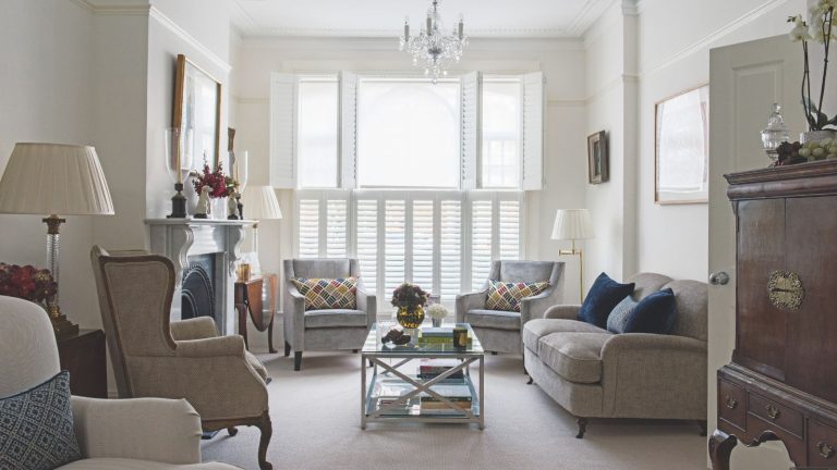 « Produit incroyable à un prix incroyable » – Les acheteurs adorent ce kit d’isolation le plus vendu à 14 $, facile à installer, pour couvrir les fenêtres en hiver A large living room with white walls, neutral upholstered couch and chairs, a glass coffee table, large mantelpiece with artwork above, and a large wooden cabinet to the right in the foreground. In the background are large windows with white shutters.