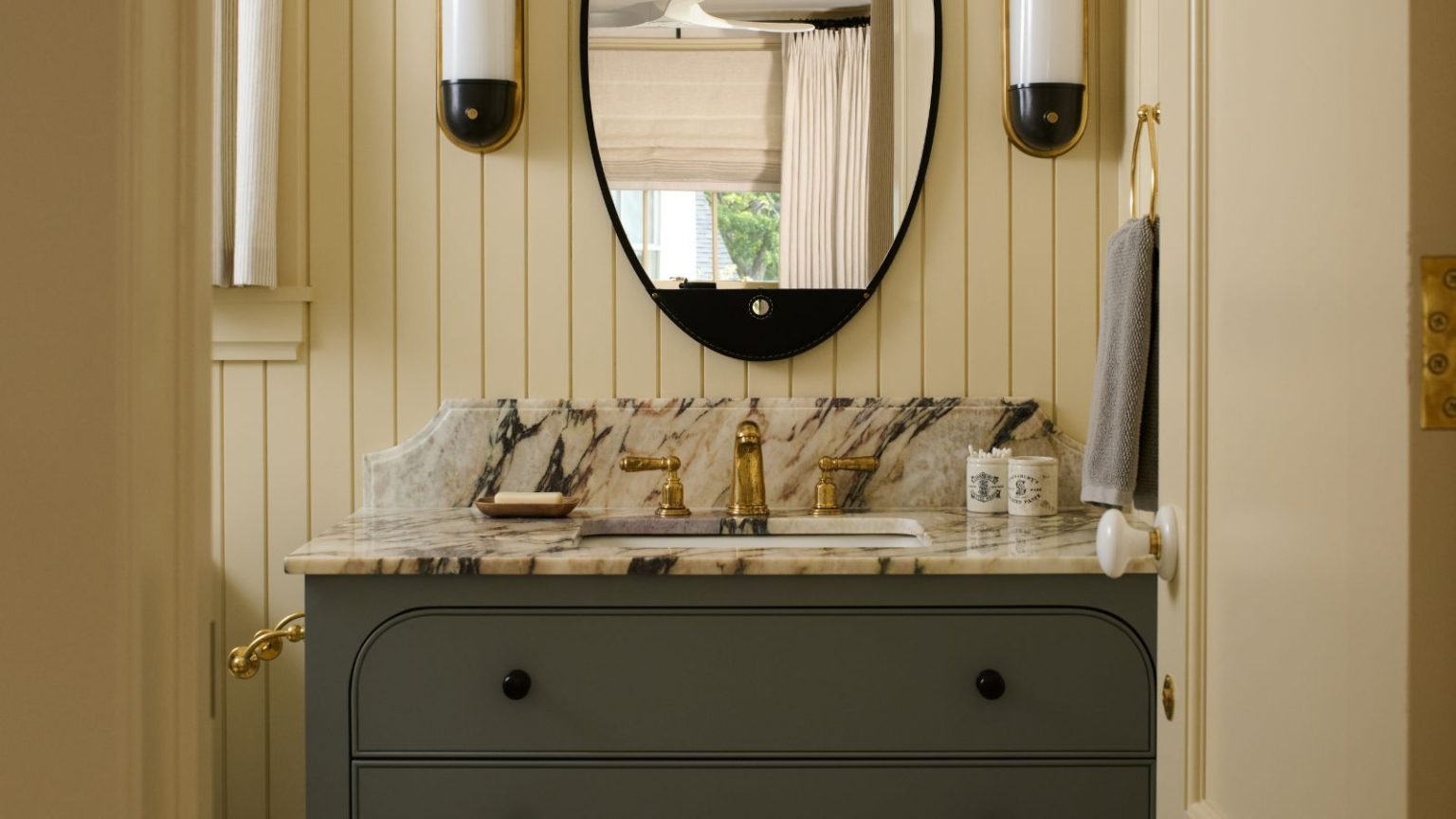 Les designers d’une seule couleur ne choisissent jamais pour les salles de bains des invités – et les teintes flatteuses qui fonctionnent toujours A butter yellow bathroom with wall panelling and a dark green cabinet with marble counters.