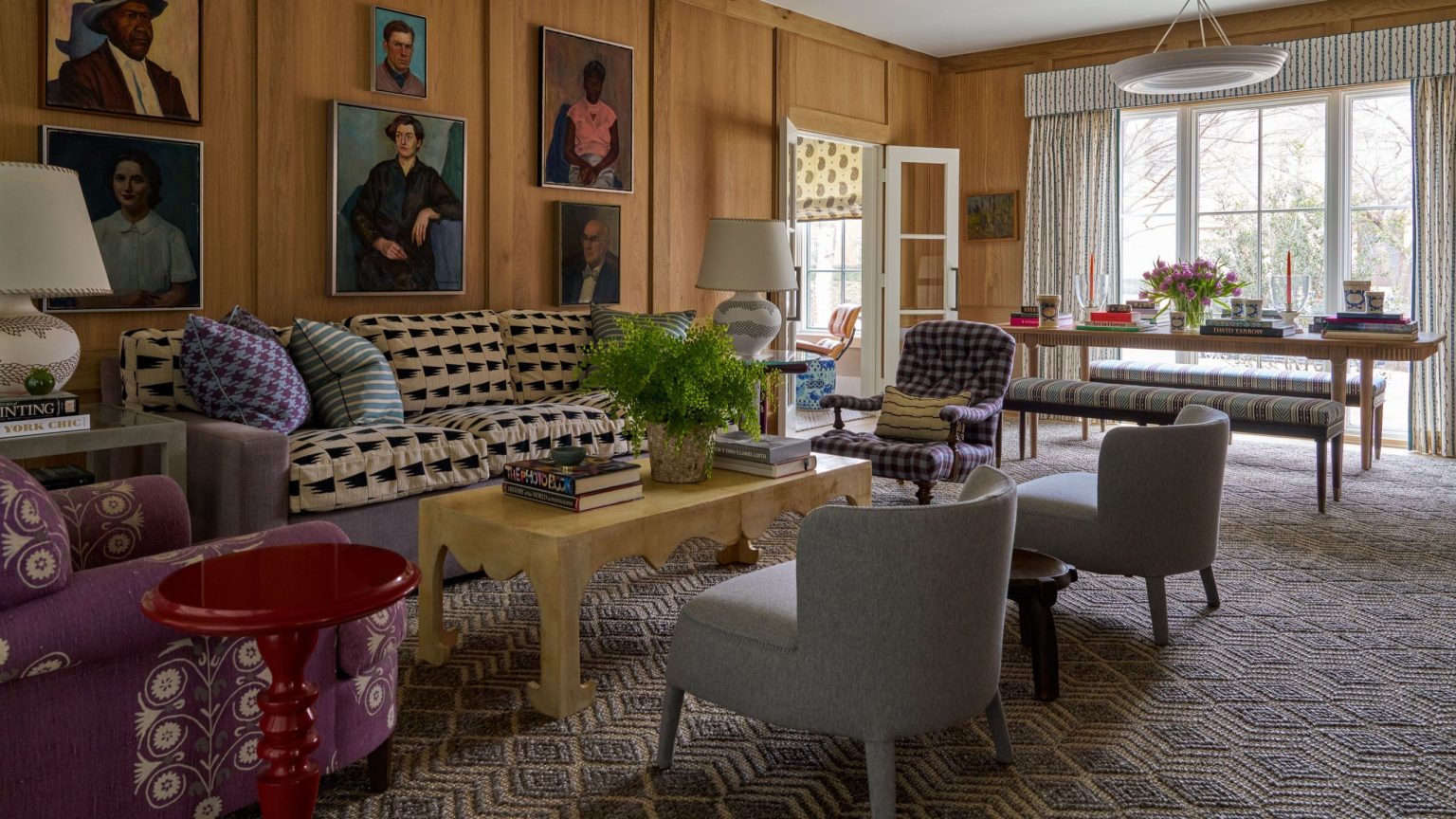 Cette maison centenaire de Dallas fait l’objet d’une rénovation ludique qui est une leçon de maximalisme silencieux A classic living room with walls fully clad in warm-toned wood paneling. A large sofa with a black and beige geometric pattern is covered in colorful pillows. Above the sofa, a collection of framed oil portraits is displayed. The room includes various patterned armchairs, a pale yellow coffee table, and a geometric-patterned rug.