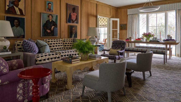 A classic living room with walls fully clad in warm-toned wood paneling. A large sofa with a black and beige geometric pattern is covered in colorful pillows. Above the sofa, a collection of framed oil portraits is displayed. The room includes various patterned armchairs, a pale yellow coffee table, and a geometric-patterned rug.