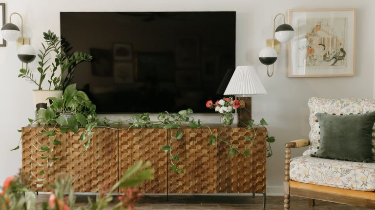 J’ai banni les « spaghettis de câble » désordonnés derrière mon téléviseur avec un simple manchon Velcro à 11 $ qui a instantanément rehaussé mon salon TV mounted above media cabinet with artificial plants running along the top. Framed artwork, a dual light fixture and a wooden armchair with cushions is pictured to the right.