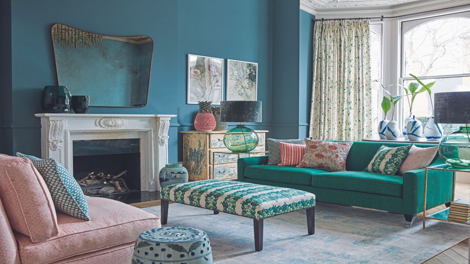 Compact, intelligent et seulement 74 $ – Le mini nettoyeur de taches Bissell qui transforme le stress lié aux taches en un jeu d’enfant A blue living room with a large white mantelpiece with a mirror above, a green sofa, cushions, a green and white ottoman, a pink sofa with cushions, and a patterned stool. To the right are large bay windows with white curtains.