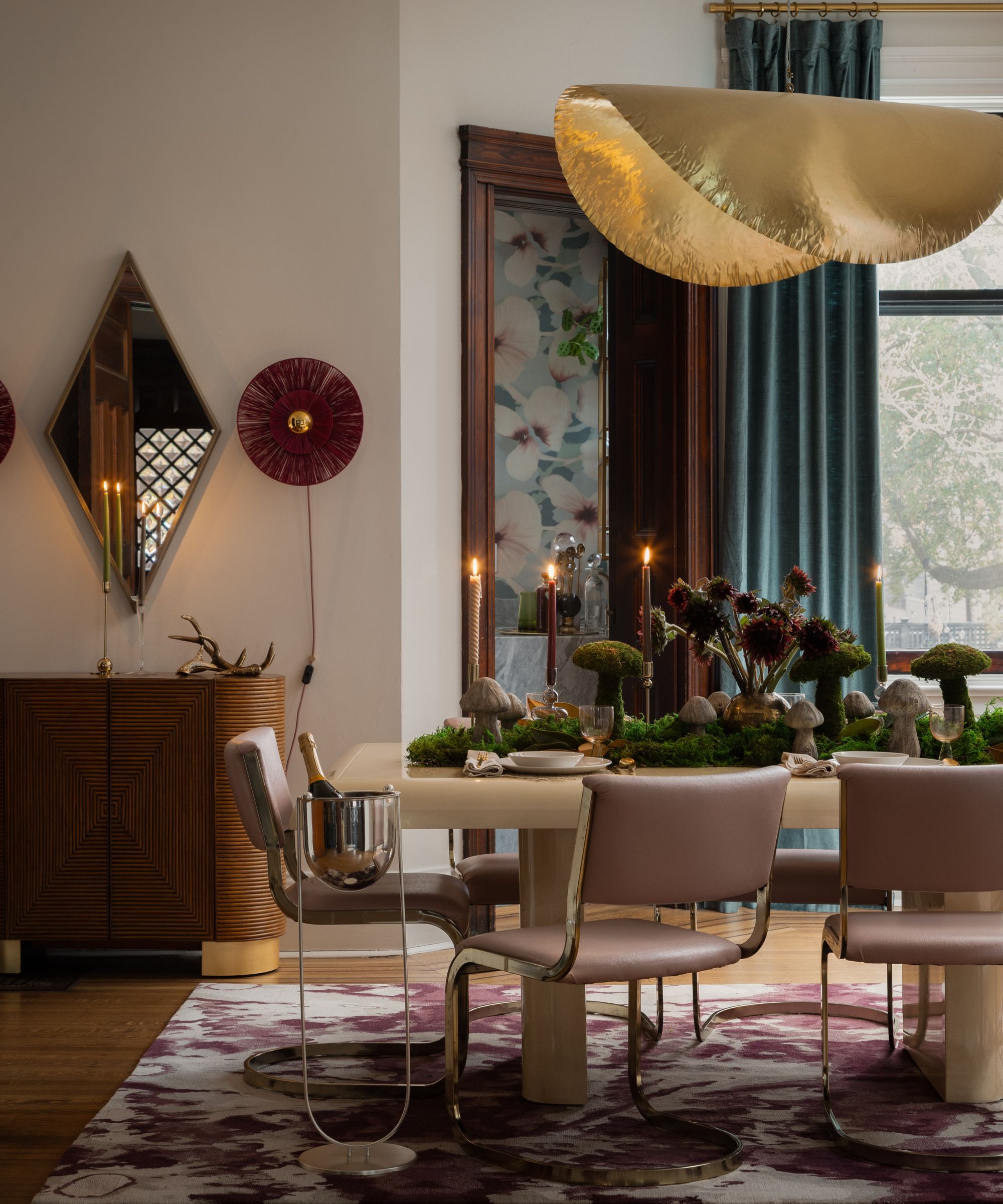 Salle à manger avec une suspension dorée ondulée, des chaises en cuir rose, une armoire en bois et une table décorée de mousse et de champignons