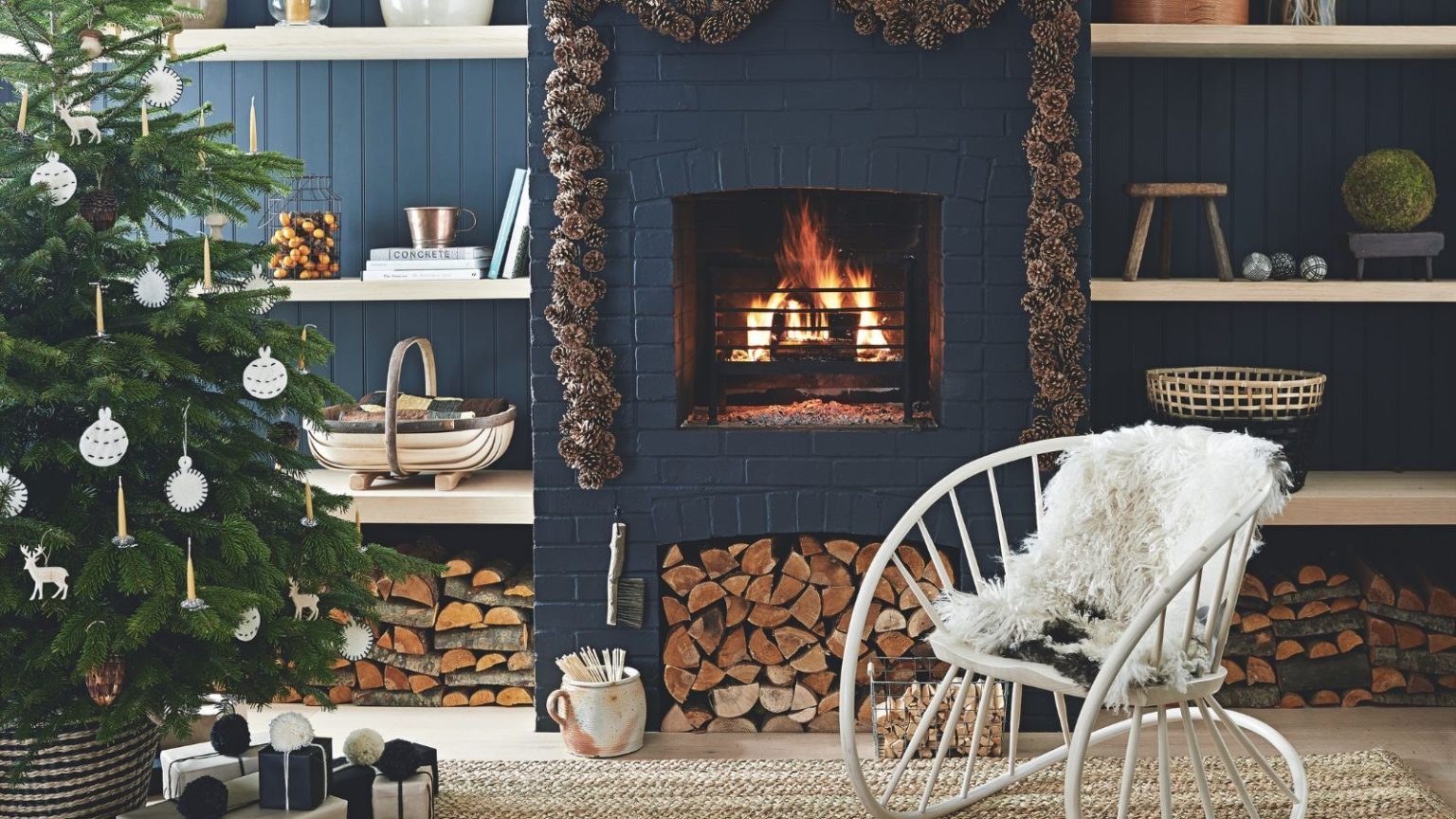 Déplacez votre arbre de Noël à cet endroit maintenant pour le bien-être et la richesse en 2026, révèlent les professionnels du Feng Shui A dark blue living room with panelled walls, shelves, and a brick fireplace with a lit fire. Around the fire is a pinecone garland, and to the left in the foreground is a large Christmas tree decorated with white ornaments. To the right of it is a large metal white statement chair with white fluffy cushion.