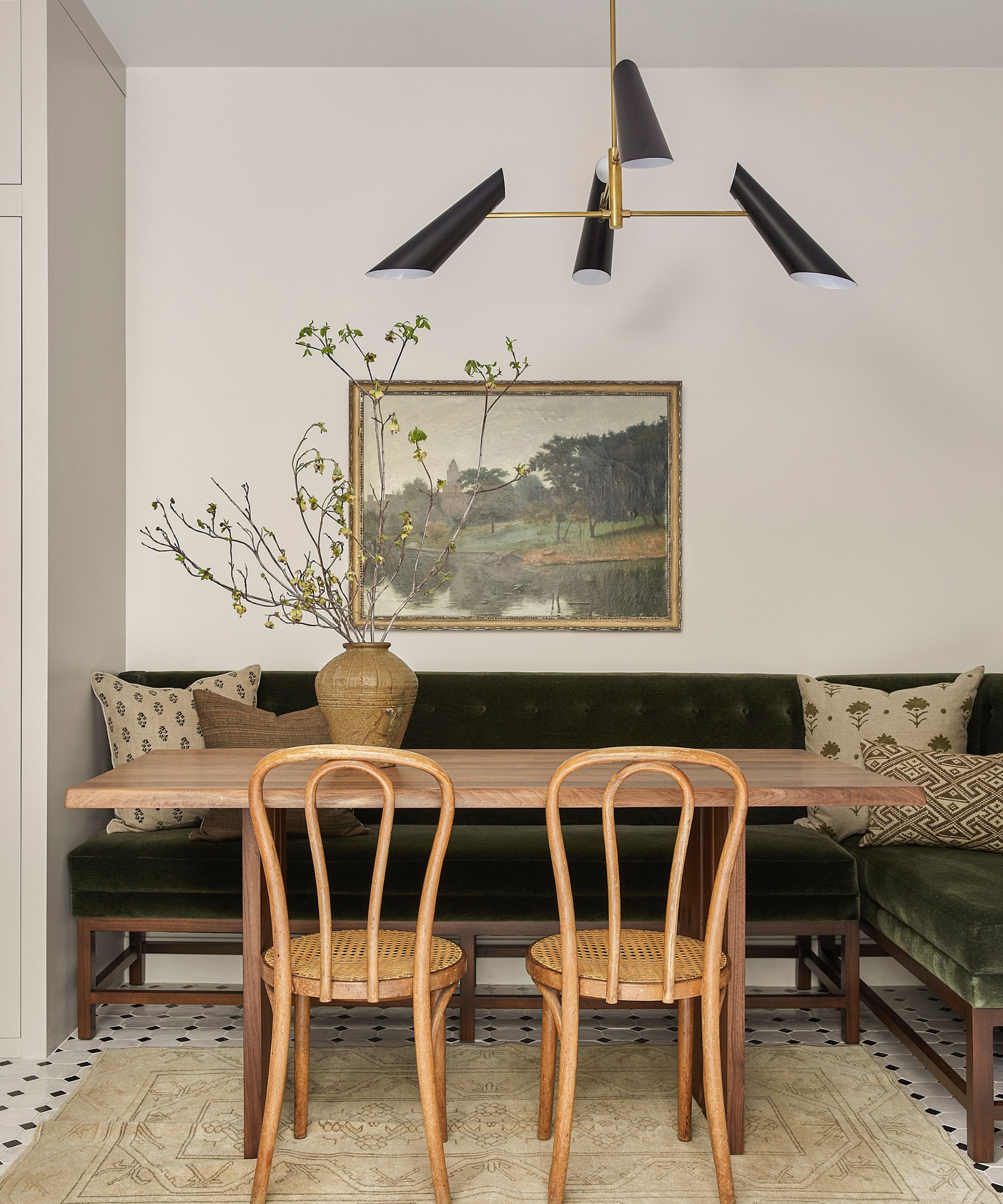 Coin repas avec une longue table en bois, une banquette en velours vert foncé, des chaises en rotin et un lustre moderne.