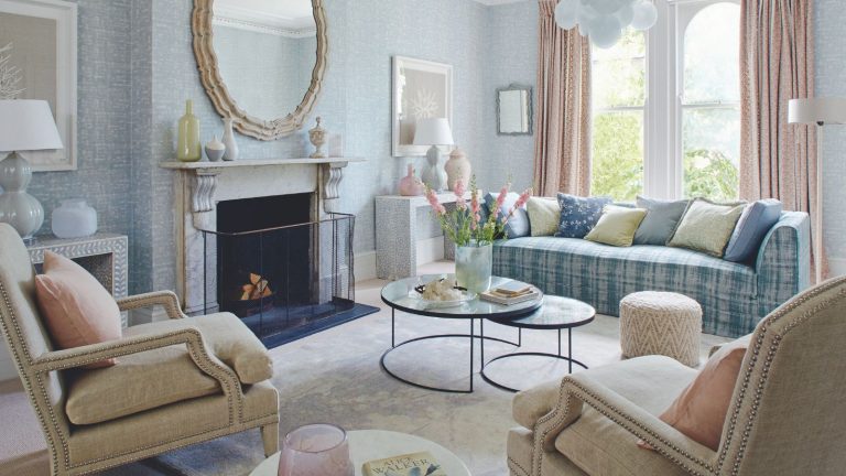 J’ai vaincu la procrastination des tâches et éliminé le désordre rapidement avec la méthode de désencombrement Domino – je suis choqué de voir à quel point cela a fonctionné A pastel living room with blue patterned wallpaper, a white marble mantelpiece with circular mirror above, and large windows to the right with pastel pink curtains. In the foreground are two cream upholstered chairs, and in the center of the room is a circular coffee table with pink flowers on it, with a blue plaid sofa behind. On the floor is a white velvet rug.