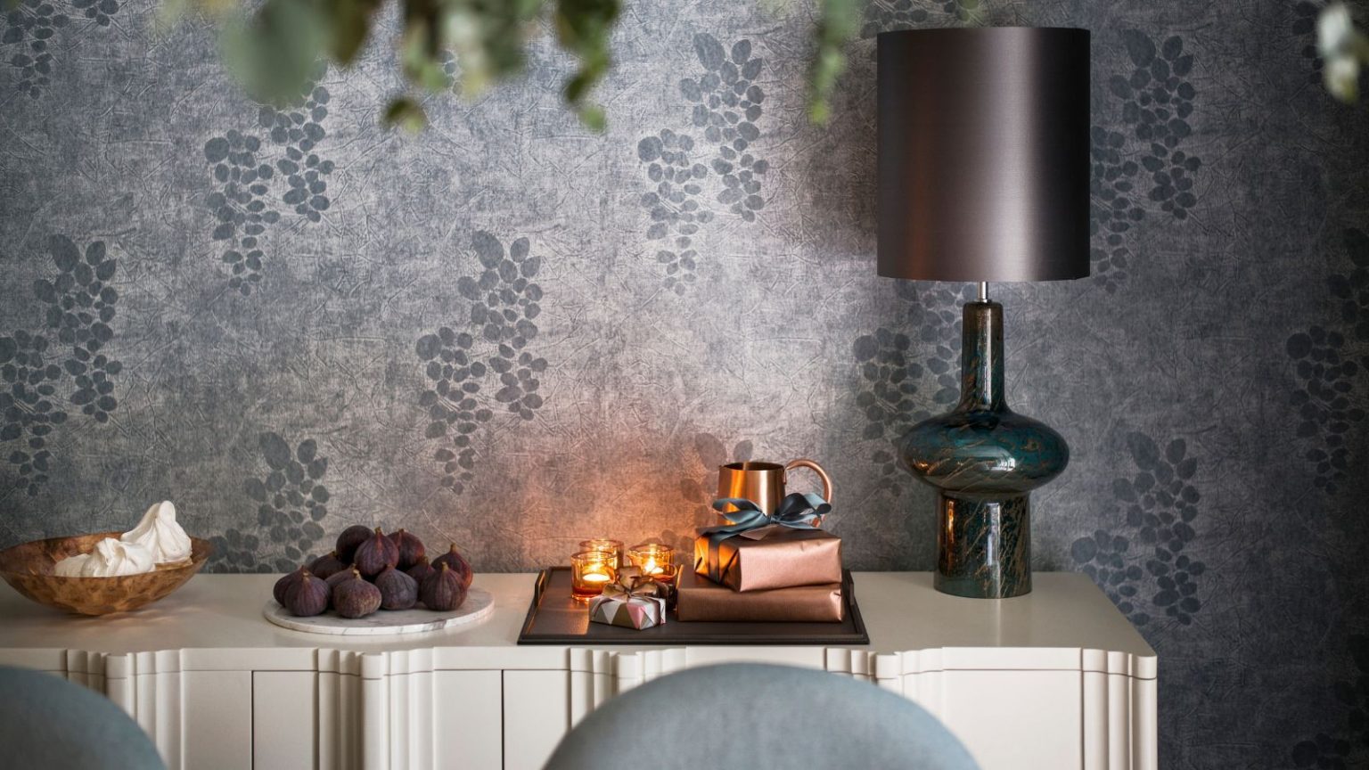 Vous stockez mal votre papier d’emballage – Ce bac de rangement à 19 $ évite les plis, les déchirures et maintient les rouleaux organisés sans effort A white side table with a wooden bowl of meringues, a white plate of figs, and a tray with tealight candles, presents wrapped in bronze paper, a small bronze jug, and a glass lamp with a grey lampshade. The walls are wallpapered with silver paper with grapes on.