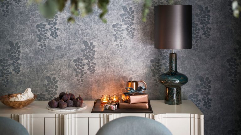 Vous stockez mal votre papier d’emballage – Ce bac de rangement à 19 $ évite les plis, les déchirures et maintient les rouleaux organisés sans effort A white side table with a wooden bowl of meringues, a white plate of figs, and a tray with tealight candles, presents wrapped in bronze paper, a small bronze jug, and a glass lamp with a grey lampshade. The walls are wallpapered with silver paper with grapes on.
