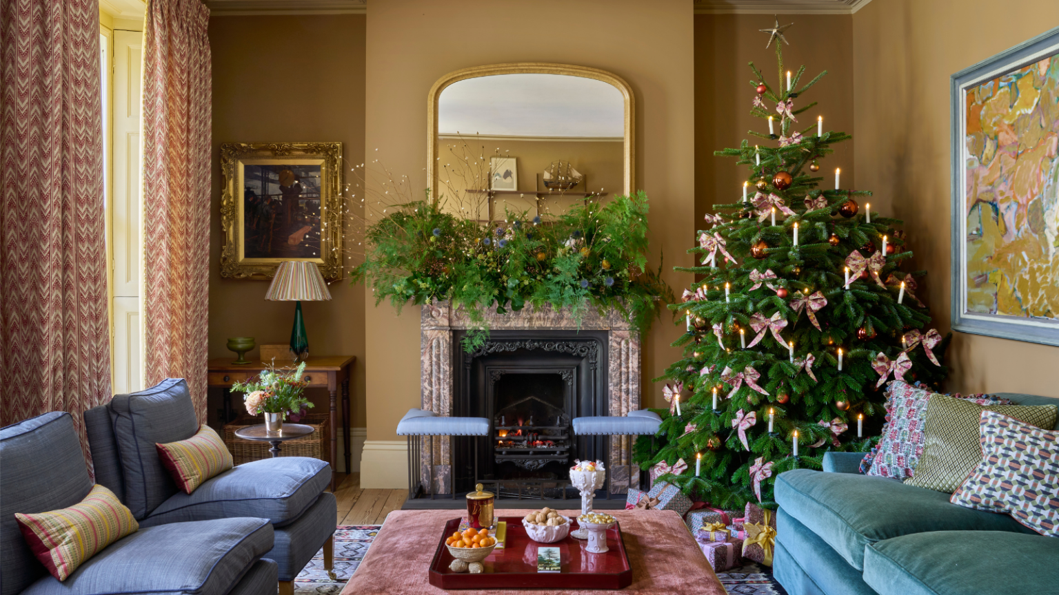 Shea McGee vient de confirmer que les nœuds sont de retour pour Noël 2025 – et elle a choisi la couleur et le style parfaits pour une décoration de vacances chaleureuse et nostalgique a living room with a teal couch, two blue accent chairs and a coffee table decorated for Christmas with a Christmas tree in one corner and garland on the mantel
