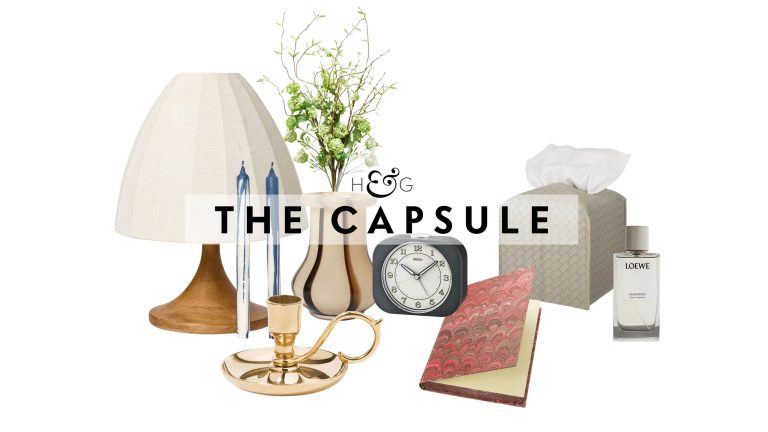 Bedside table decor pictured against a white background, including a table lamp, taper candle duo, brass candlestick, striped vase, retro-style alarm clock, patterned notebook, woven leather tissue cover, and Loewe Home spray