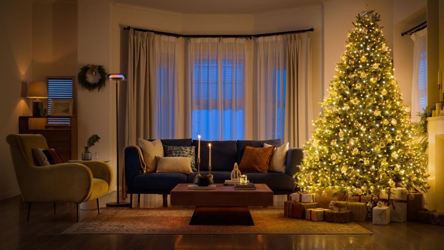 L’experte en design Genevieve Gorder explique comment transformer votre maison en une retraite enchanteresse avec un éclairage de Noël sans effort Living room with large windows, navy blue sofa, armchair and large christmas tree with warm lights