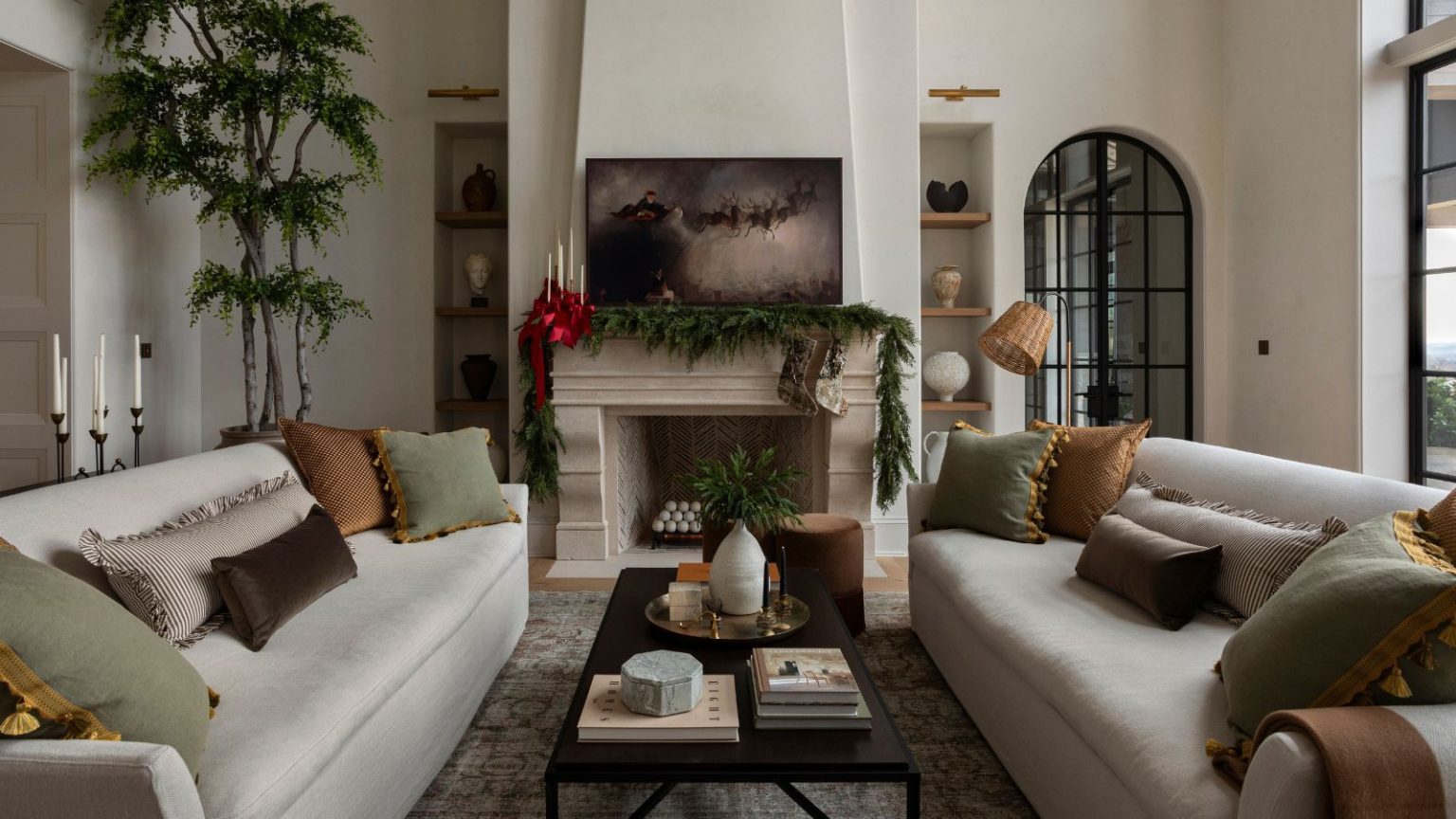 La designer Lauren Sullivan affirme que la clé d’une décoration de vacances raffinée est de choisir un seul détail caractéristique – voici comment suivre ses conseils bright living room with two white sofas facing a stone fireplace decorated with evergreen garland, red ribbons, and a painting of Santa and his sleigh