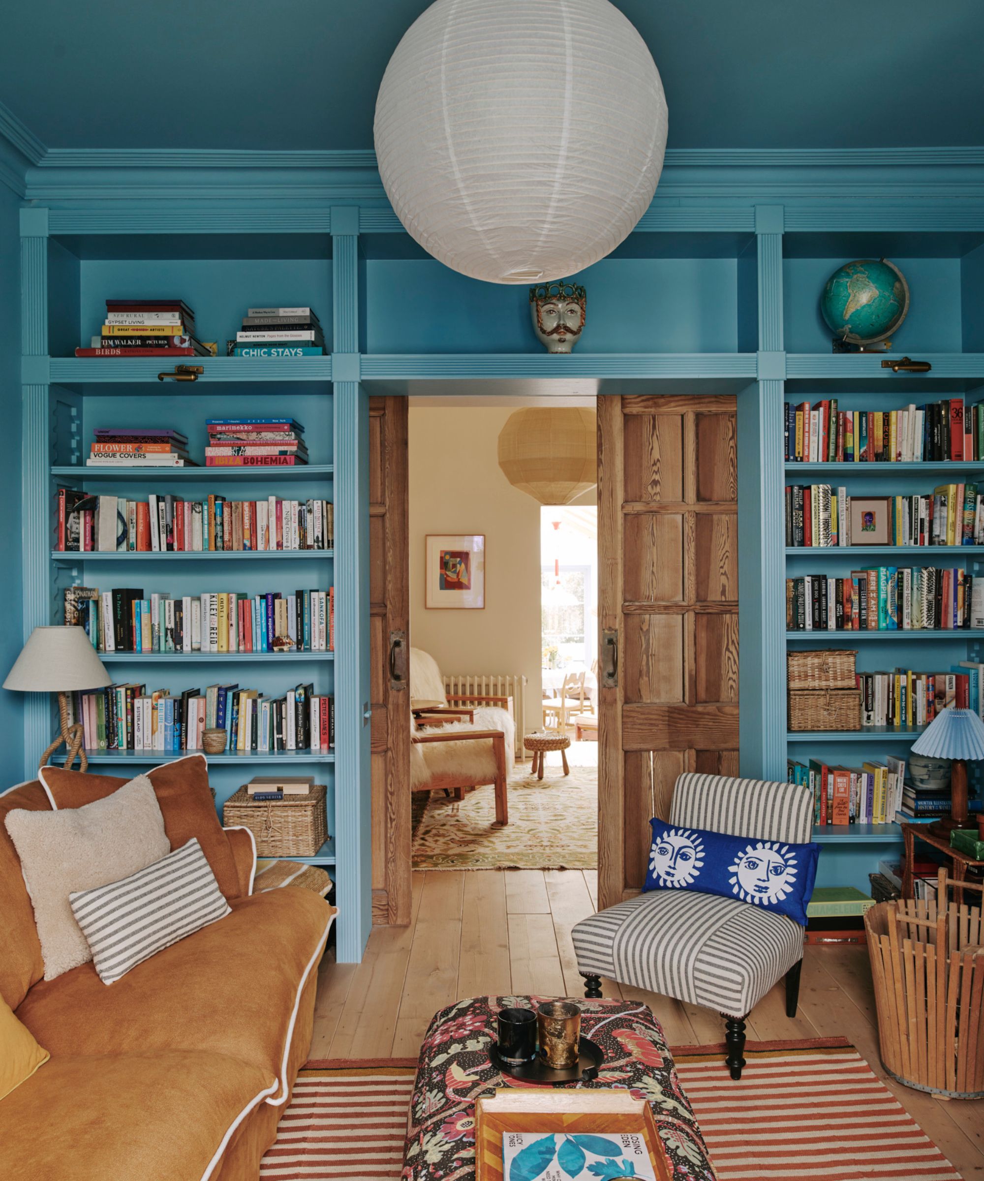 Un salon confortable avec des murs de couleur bleue avec des étagères intégrées, un canapé marron, un tapis à rayures et un pouf à motifs.