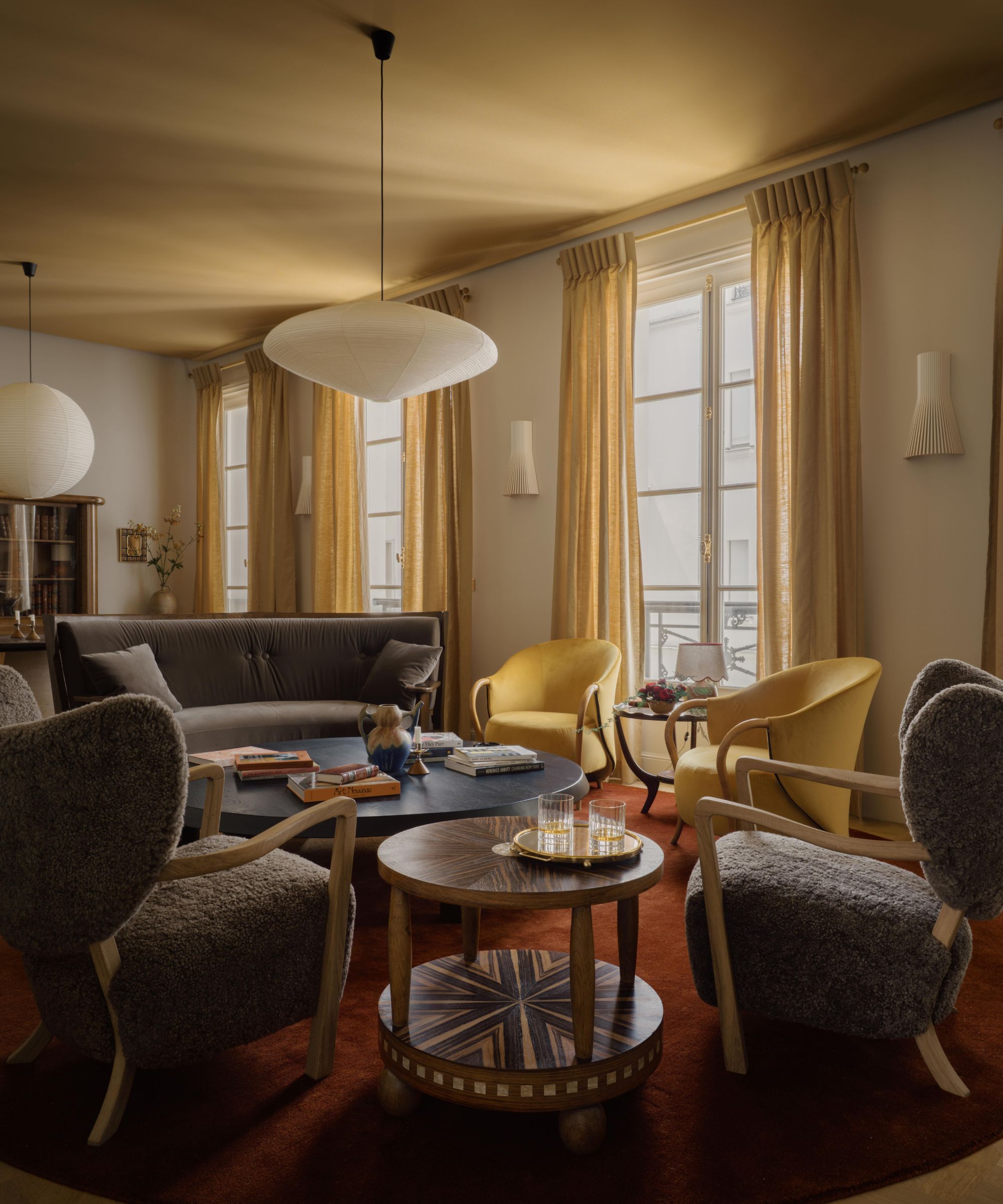 Un salon avec un plafond ocre, des murs blanc chaud et une grande fenêtre avec des rideaux ocre. Un tapis rouge foncé et des chaises sombres et neutres.