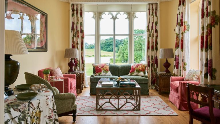 A living room filled with layered patterns with wooden flooring and a large gothic window with views to the garden. There are red and green upholstered chairs and a small sofa ,and floral curtains and cushions.