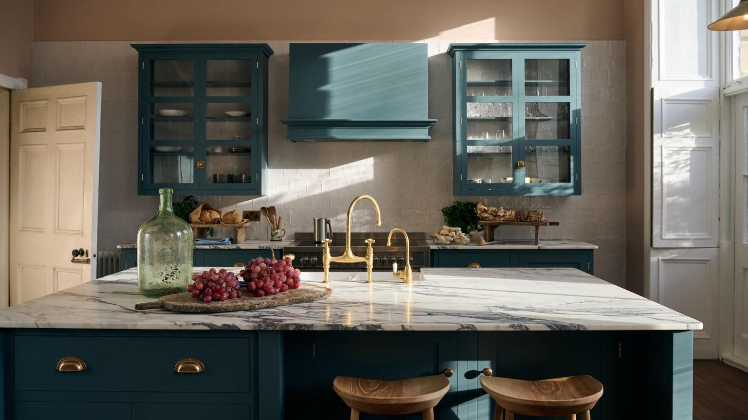 Cette astuce simple avec de l’eau chaude et un couteau à citron réduit de moitié le temps de nettoyage lors de la cuisson de gros repas A kitchen with muted blue cabinets, large island with gold faucet, wooden chairs, fruit on wooden board and white veined gray marble countertops