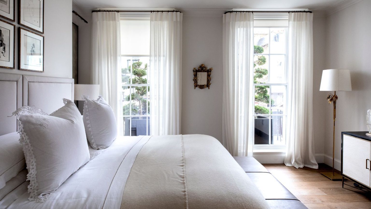 Pourquoi vos murs blancs semblent jaunes – et les nuances de peinture parfaites que les experts en couleurs choisissent pour y remédier A white bedroom with a double bed with white bedding, white walls, wooden flooring, a gold lamp with white lampshade, black and white cabinetry, and two large windows with sheer white curtains.