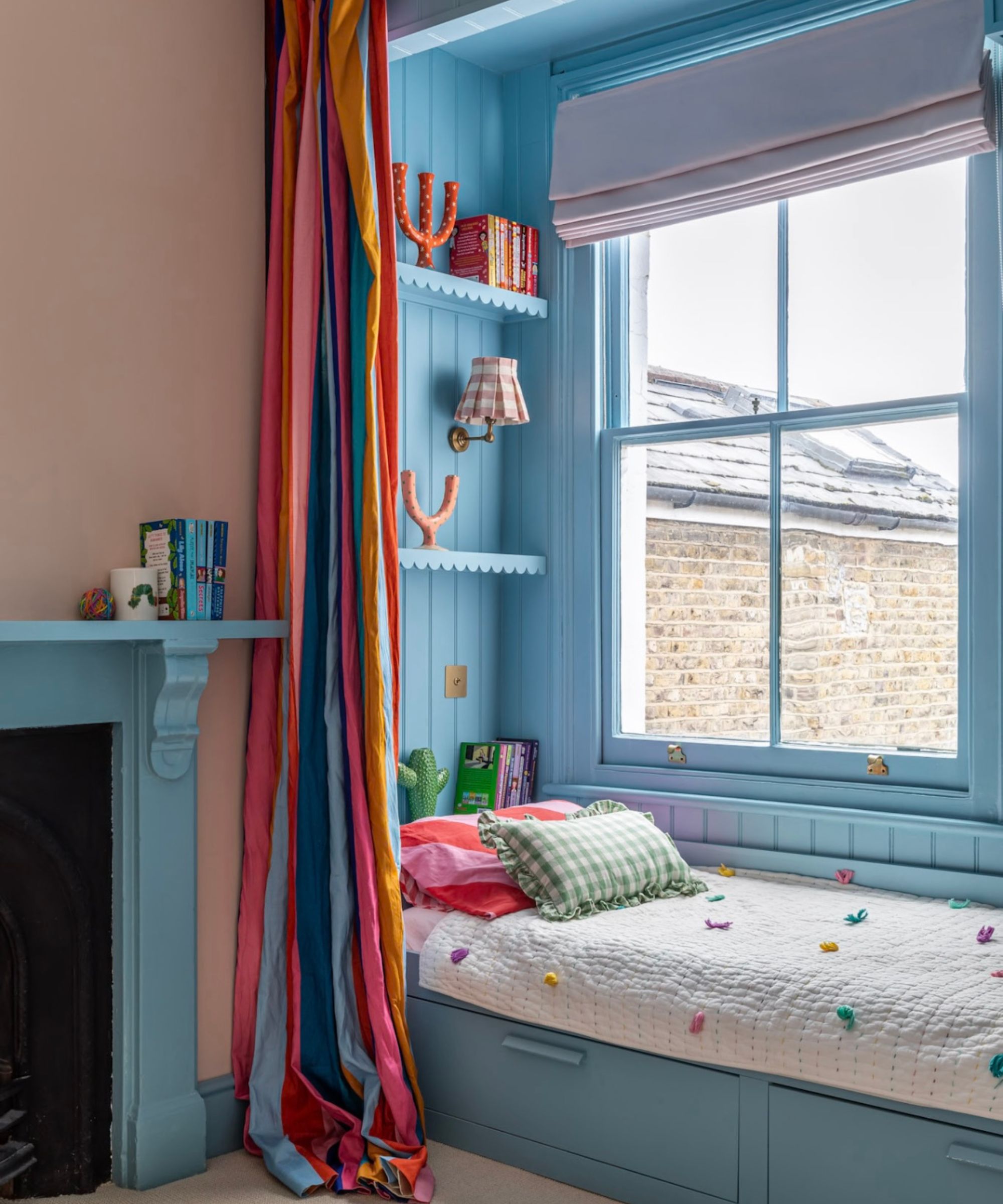 Une chambre d'enfant avec des murs en plâtre rose, un lit cabane bleu à côté d'une fenêtre avec un rideau coloré et une cheminée bleue.