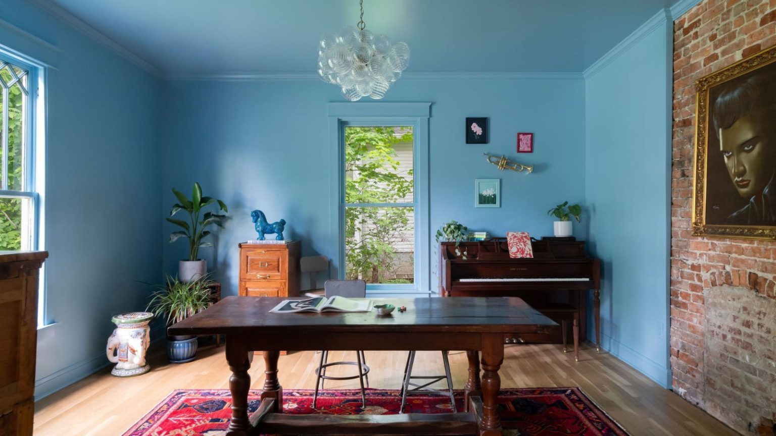 Cette teinte exacte de Farrow & Ball est la couleur du moment, mais sa popularité durera-t-elle ? Les créateurs se prononcent sur l’emblématique bleu poudre « Yonder » A parlor painted blue with a large wooden desk, a red rug, and a piano in the corner.