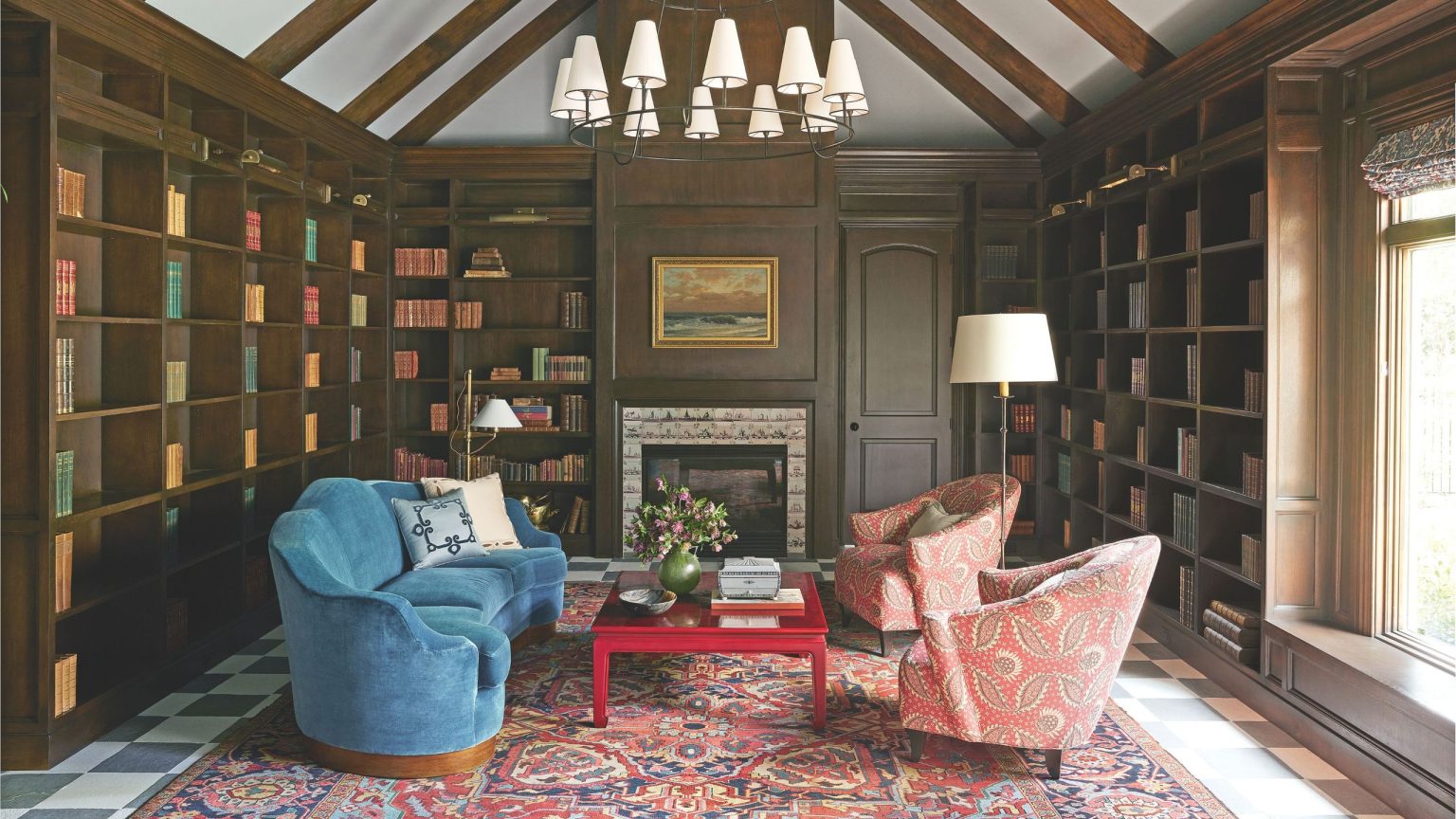 Cette maison est peut-être située au cœur du Texas, mais son inspiration vient de la pittoresque campagne anglaise A grand, wood-paneled home library with floor-to-ceiling bookshelves, a vaulted beamed ceiling, a large tiered chandelier, and a plush blue sofa facing patterned coral armchairs over a Persian rug