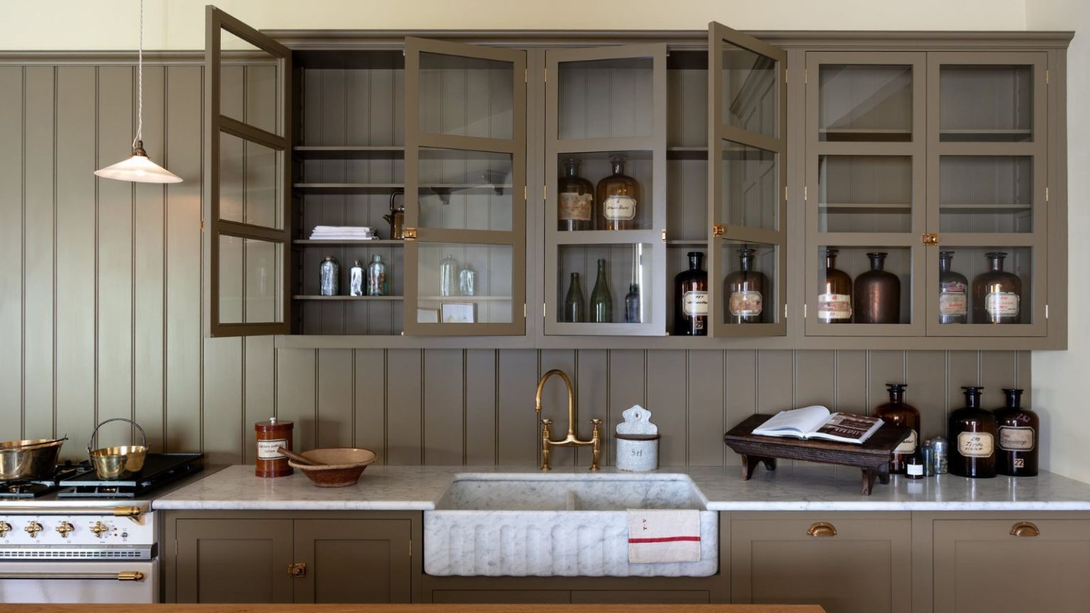 5 choses à ne jamais ranger au-dessus de vos armoires de cuisine – et que faire avec elles à la place Taupe kitchen cabinets with glass fronts, butler sink, brass fixtures, and paneled walls. There are amber glass jars and a cookbook in view