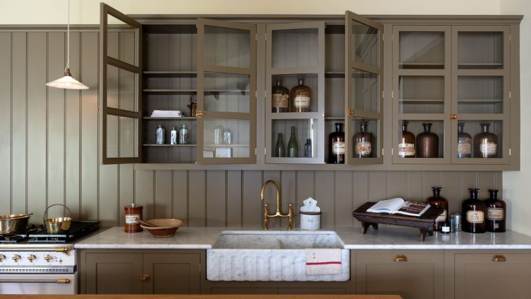 5 choses à ne jamais ranger au-dessus de vos armoires de cuisine – et que faire avec elles à la place Taupe kitchen cabinets with glass fronts, butler sink, brass fixtures, and paneled walls. There are amber glass jars and a cookbook in view