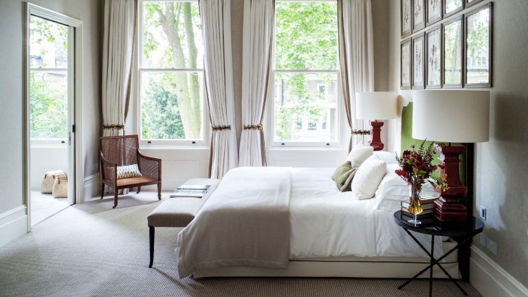 A bright and white carpeted minimalist bedroom with a large bed with white and beige bedding and green headboard, black circular bedside tables, red lamps with white lampshades, a framed gallery wall and a brown wicker chair.