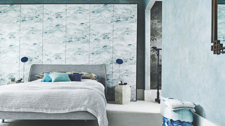 A blue bedroom with a grey double bed, statement wallpaper wall, two matching blue lamps and a blue patterned stool.