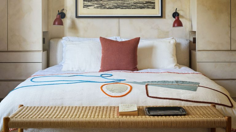 L’édition très britannique : comment j’utilise le calme de janvier pour transformer ma maison en un espace qui nourrit, restaure et rend la vie quotidienne plus heureuse A brightly lit, modern bedroom featuring a bed with a white textured duvet decorated with abstract, colorful embroidered shapes in blue, orange, and green. At the foot of the bed sits a woven light-wood bench holding a book and a black tray. The headboard is flanked by built-in light wood cabinets and two small red wall-mounted sconces. Above the bed hangs a large framed horizontal piece of art.