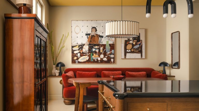 Vous aimez les couleurs terreuses mais vous ne voulez pas que votre maison ait l’air terne ? Découvrez la tendance montante de 2026 : « Earthy Vibrancy » A kitchen with warm white walls, a wooden island and dresser, a red velvet sofa and a yellow ceiling.
