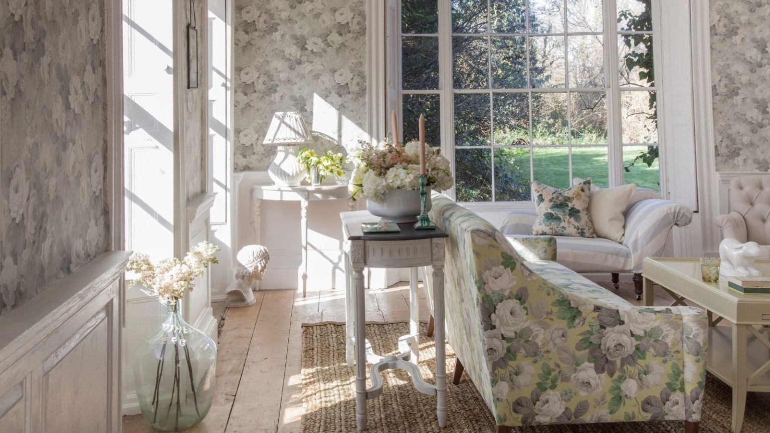 3 tendances de nettoyage à faible effort que nos rédacteurs en chef tentent d’économiser du temps et de l’énergie en 2026 A floral living room with floral wallpaper, floral couch, white coffee table, rattan rug, wooden flooring, large glass vase with white flowers, and large windows out onto a leafy garden.