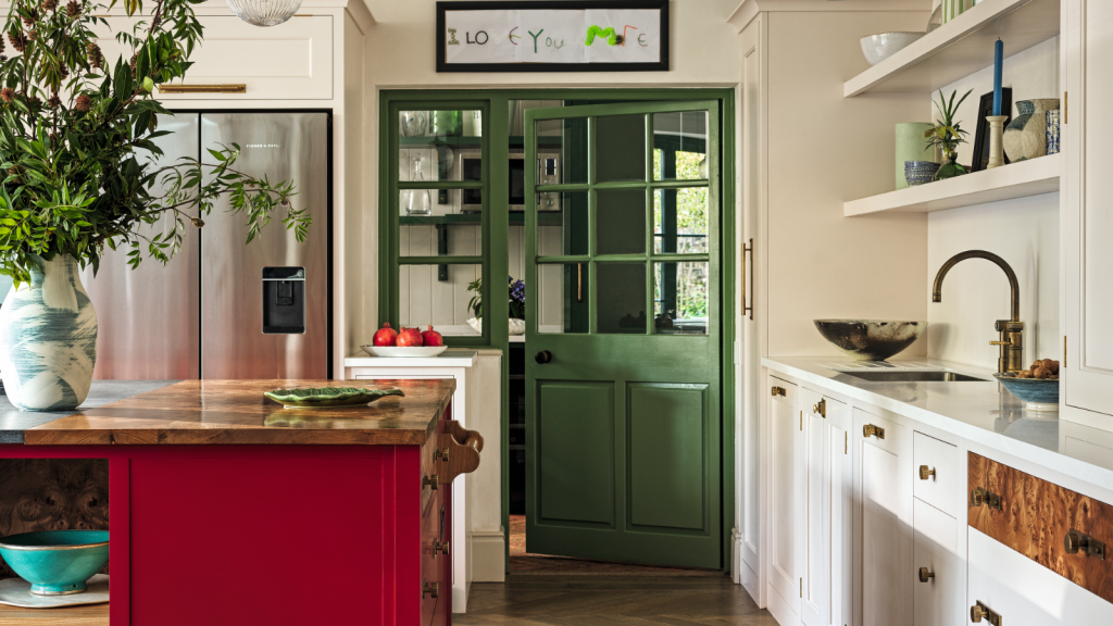 Les portes d’accent sont la réponse de 2026 au mur d’accent obsolète – une façon subtile et sophistiquée d’ajouter de la couleur instantanée a kitchen with a red kitchen island, white cabinets, open shelves, and a green pantry door