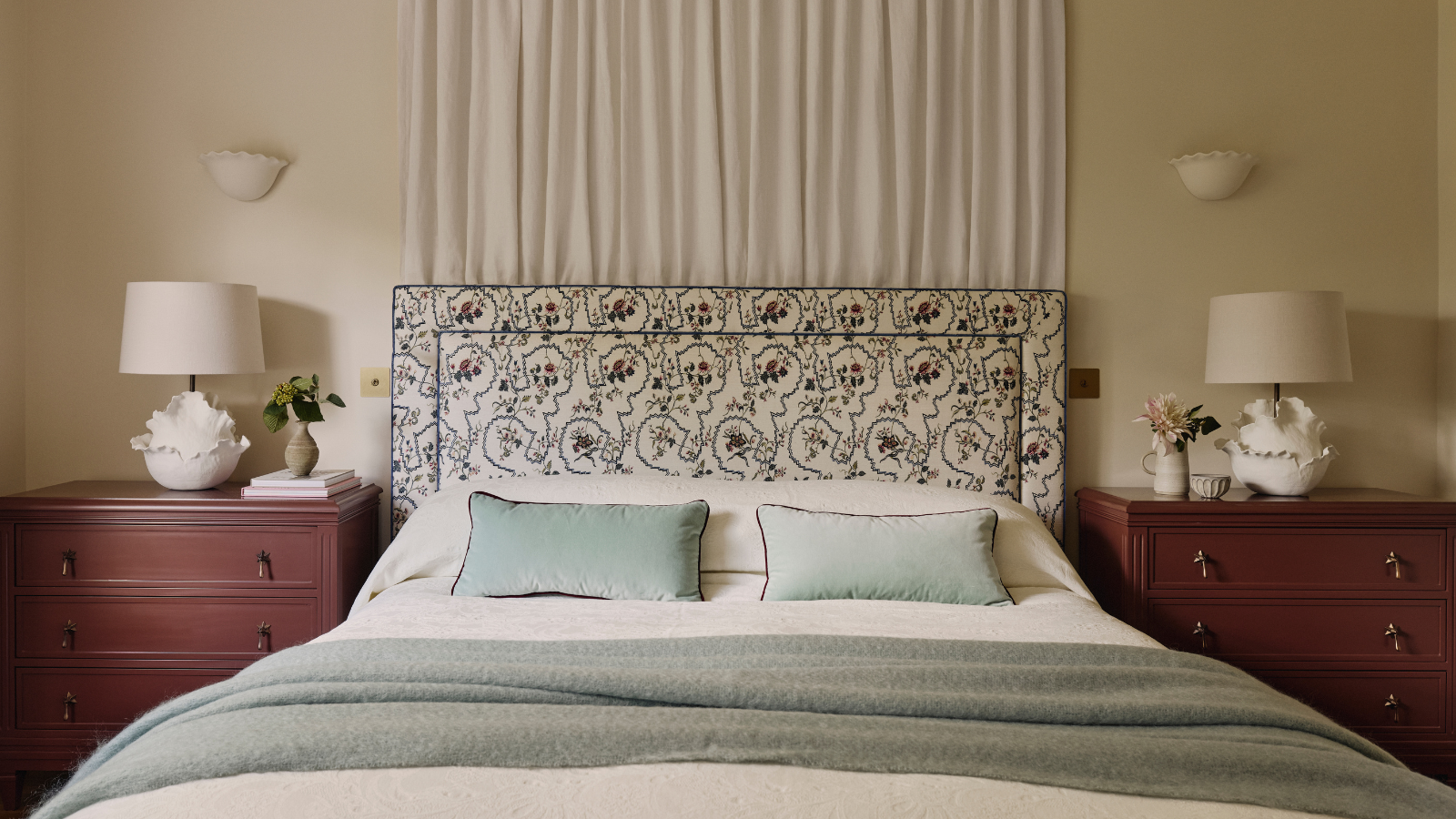 un lit avec une tête de lit fleurie, deux commodes de chaque côté avec des lampes sur le dessus, des oreillers verts et un rideau derrière le lit