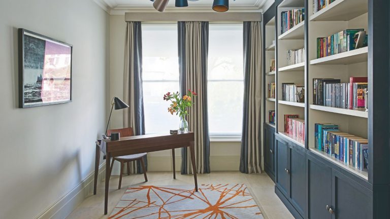 An uncluttered home office with a wooden desk, artwork, statement rug and two large windows with curtains. To the right are built-in blue and white shelves and cabinets.