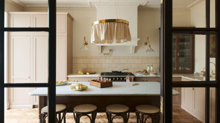 Malheureusement, la chaleur n’a jamais atteint mon salon – 1 conseil surprenant de pro CVC pour uniformiser la température en quelques minutes Character peachy pink kitchen viewed through internal black Crittal doors. There is a marble island table with wooden backless bar stools in view. In the background, there is a splashback tiled stove area and built-in pink, and glass front brown cabinets.