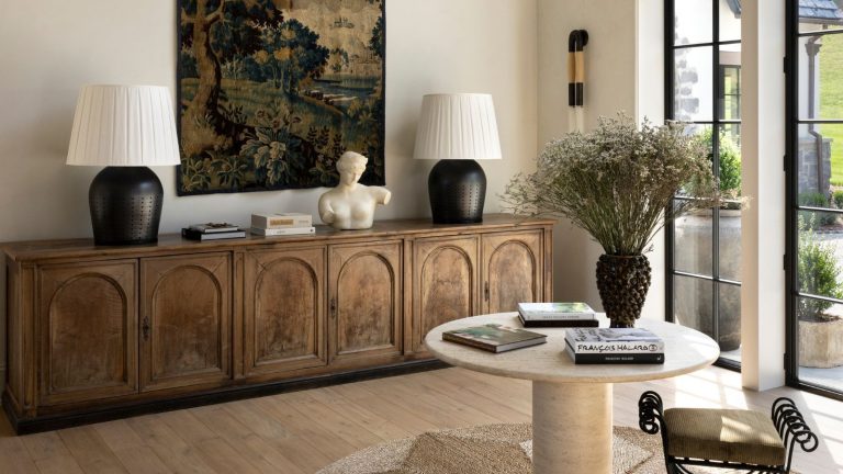 Si vous investissez dans une pièce antique pour votre maison en 2026 – les designers disent : faites-en celle-ci light bright neutral entryway with light walls and floor with a large rustic credenza sitting beneath a large antique tapestry